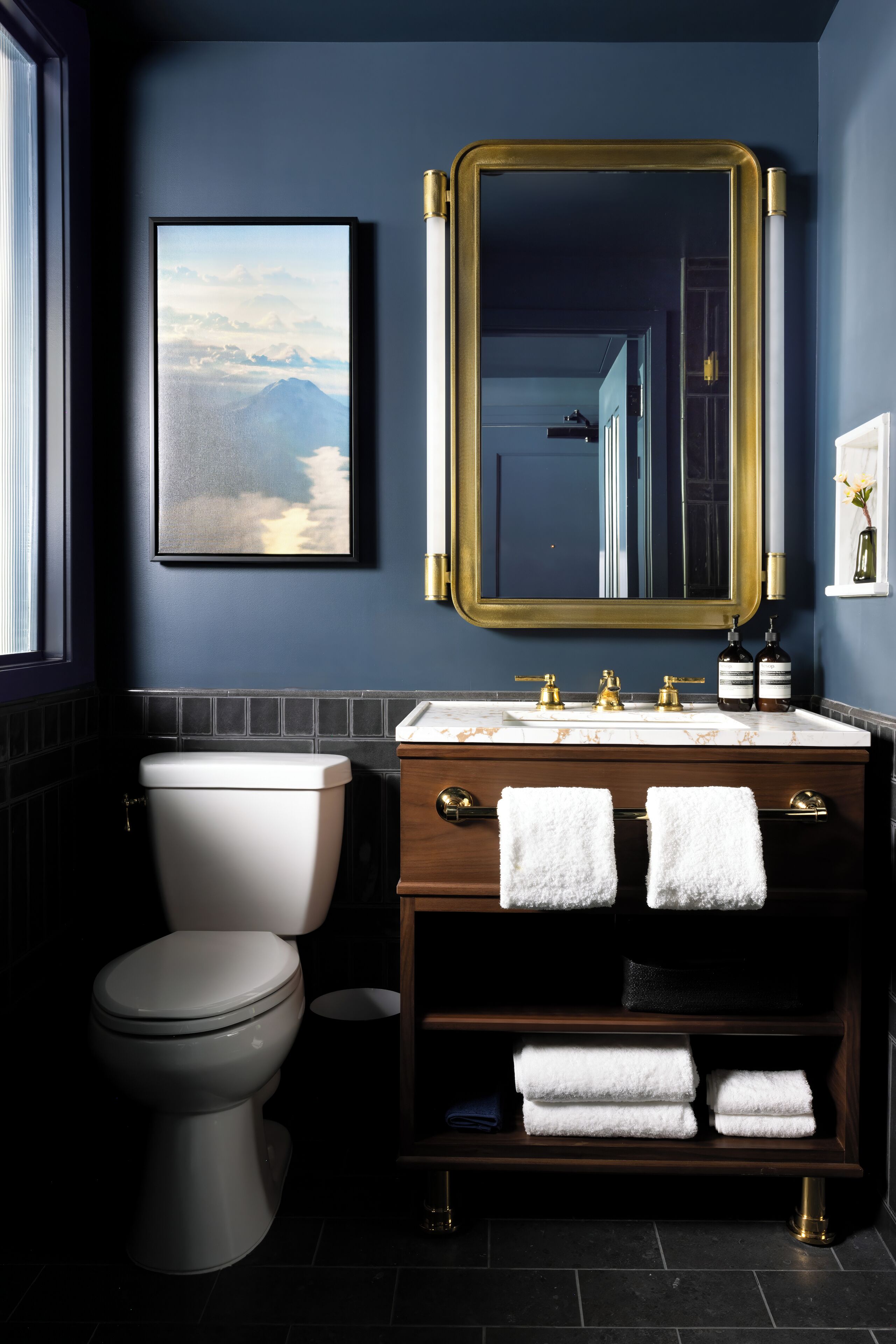 Un baño moderno con una pared azul oscuro, un cuadro de paisaje enmarcado, un espejo con marco dorado, un inodoro blanco, un lavabo de mármol y dos toallas sobre el mueble.