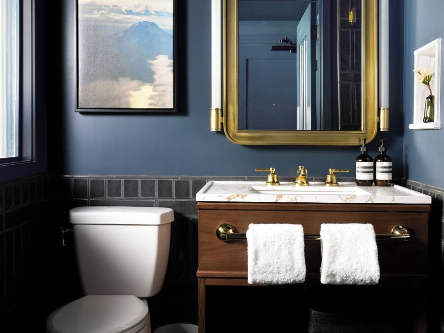 Un baño moderno con una pared azul oscuro, un cuadro de paisaje enmarcado, un espejo con marco dorado, un inodoro blanco, un lavabo de mármol y dos toallas sobre el mueble.