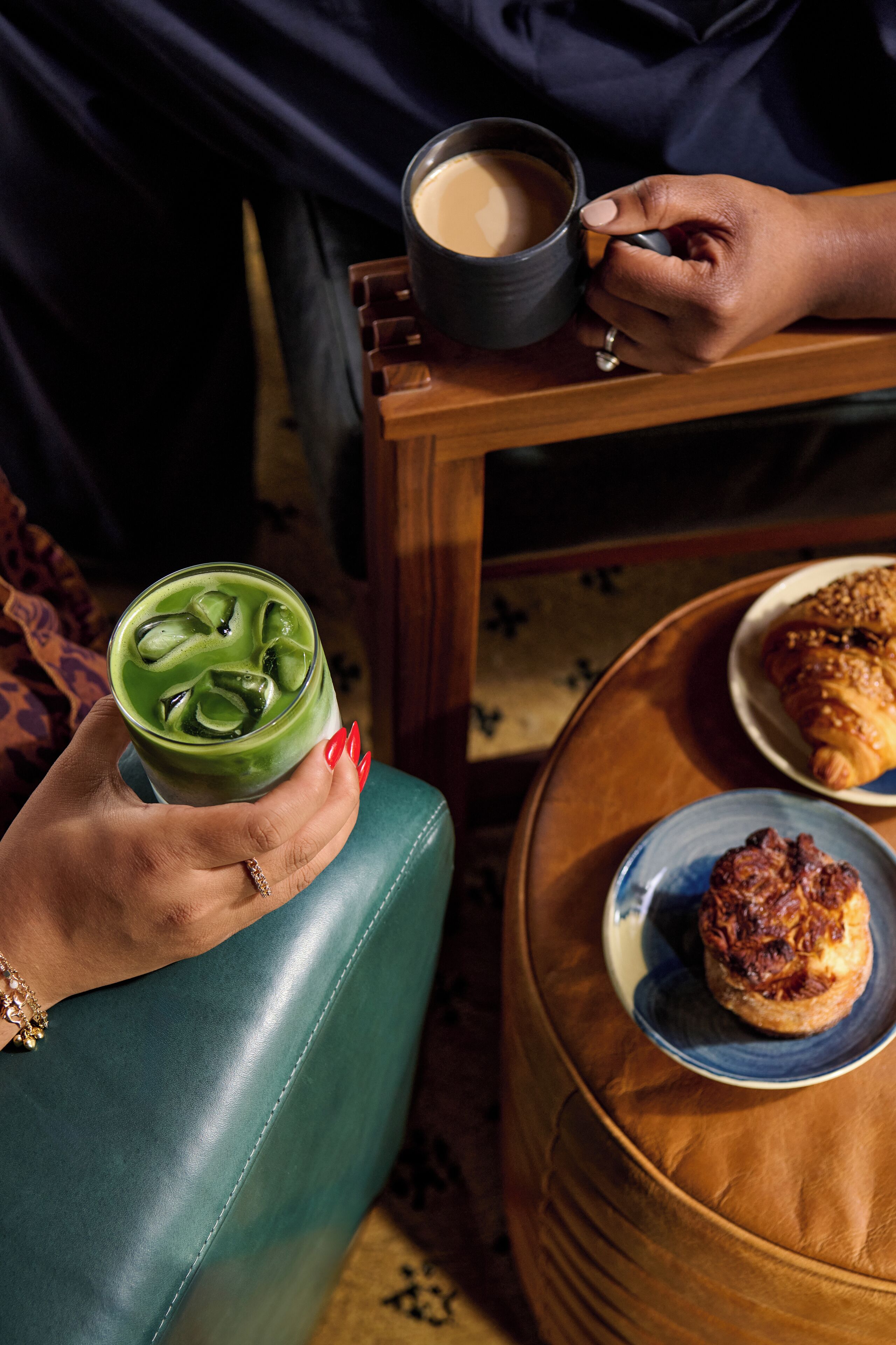 Una persona sosteniendo una bebida verde con cubitos de hielo, una taza de café y un plato con pasteles sobre una mesa de madera.