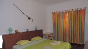 Habitación de hotel con cabecera de madera, cama con ropa de cama verde, mesita de noche con lámpara y cortina a rayas y colores.