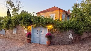 A stone building with a blue door and two potted plants, surrounded by greenery. - Assos Mia Konaklama ve Restaurant (Ayvacik)