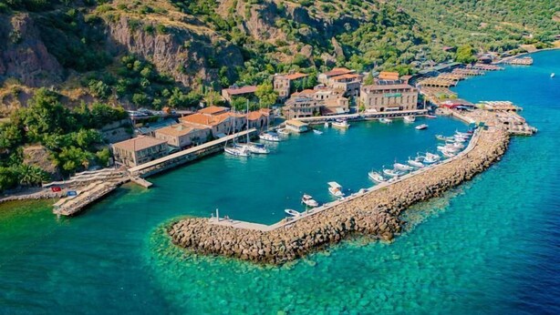 An aerial view of a coastal town with a marina, stone buildings, and clear turquoise waters. - Assos Mia Konaklama ve Restaurant (Ayvacik)