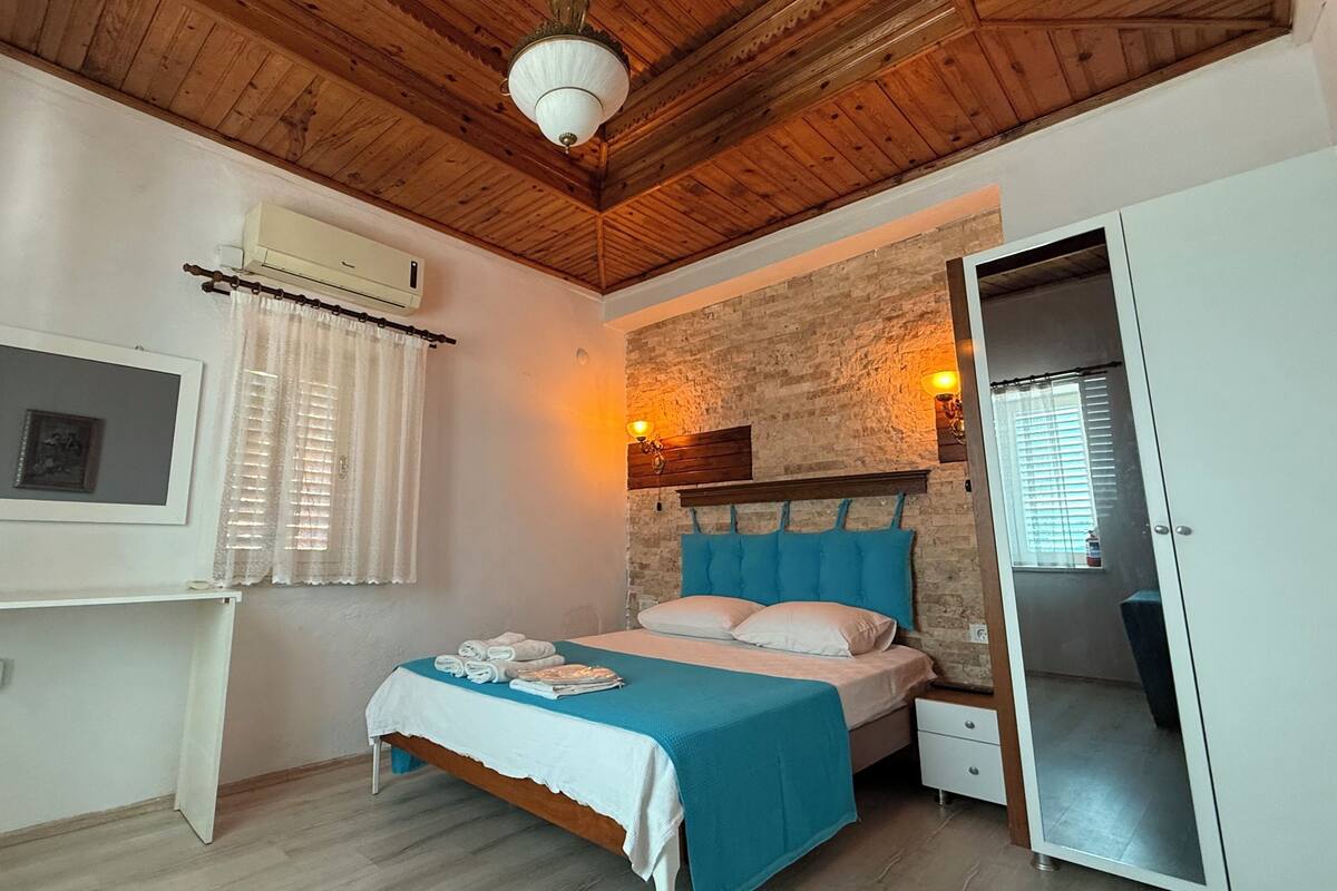 A bedroom with a bed, a nightstand, a wall-mounted air conditioner, a window with curtains, a wall-mounted light fixture, and a mirror.