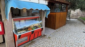 Un pequeño quiosco al aire libre con un expositor de vidrio lleno de bebidas, un toldo azul y una estructura de madera cerca.