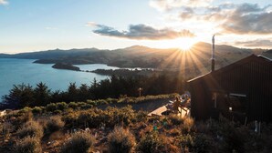 A coastal view with a wooden building, a wooden deck, and a person sitting on a chair. - Kūkupa Matairangi at Akaroa Lavender (Duvauchelle)