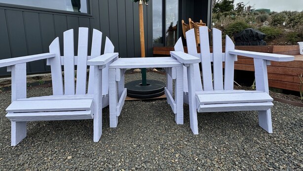 Two white Adirondack chairs and a small table set on gravel outside a building with a green umbrella. - Kūkupa Matairangi at Akaroa Lavender (Duvauchelle)