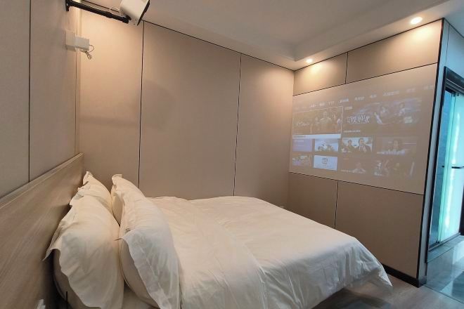 Quarto de hotel moderno com cama, projetor fixado na parede e uma janela grande.