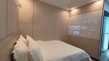 Quarto de hotel moderno com cama, projetor fixado na parede e uma janela grande.