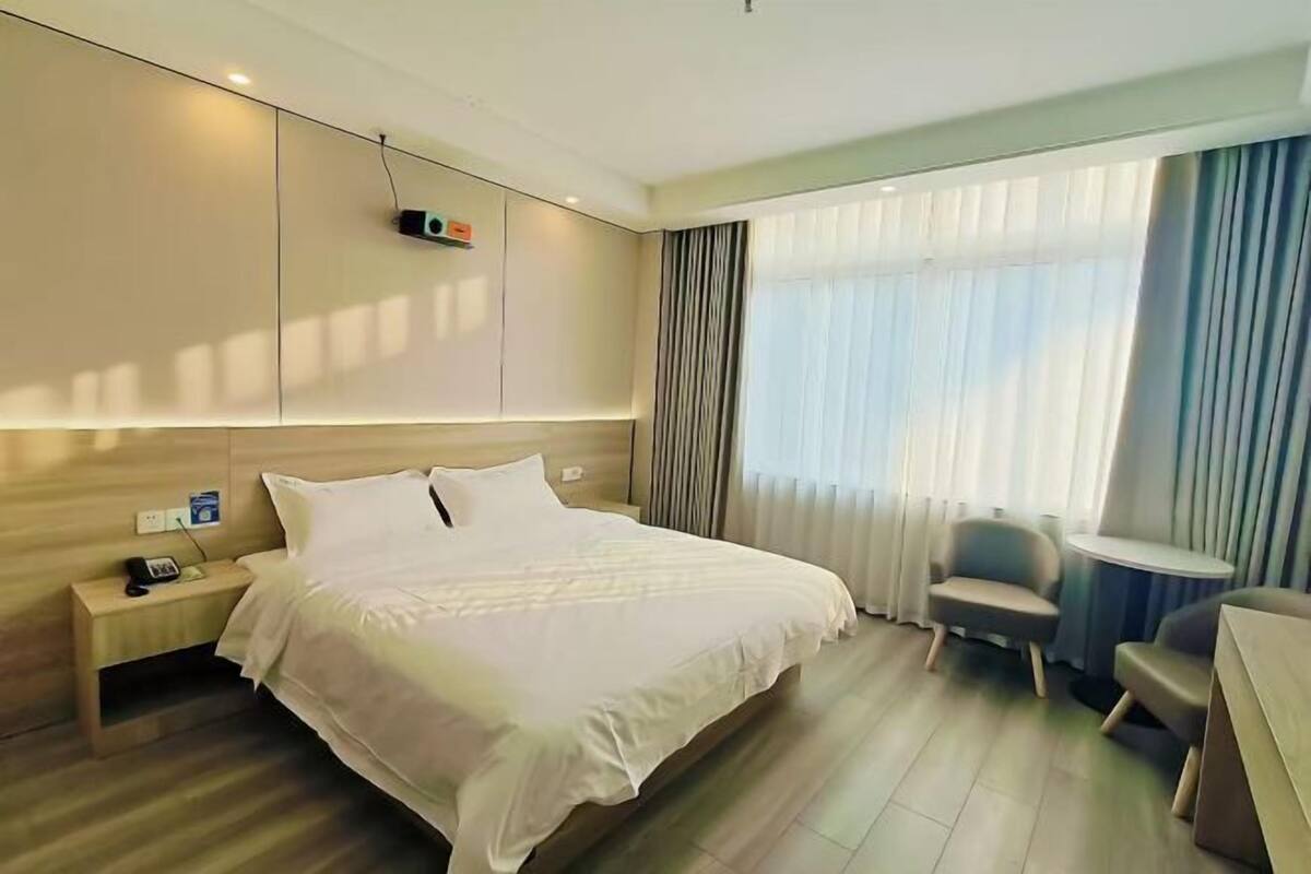 Ein Hotelzimmer mit Bett, Nachttisch, Lampe, Stuhl und Blick ins Freie durch ein Fenster.