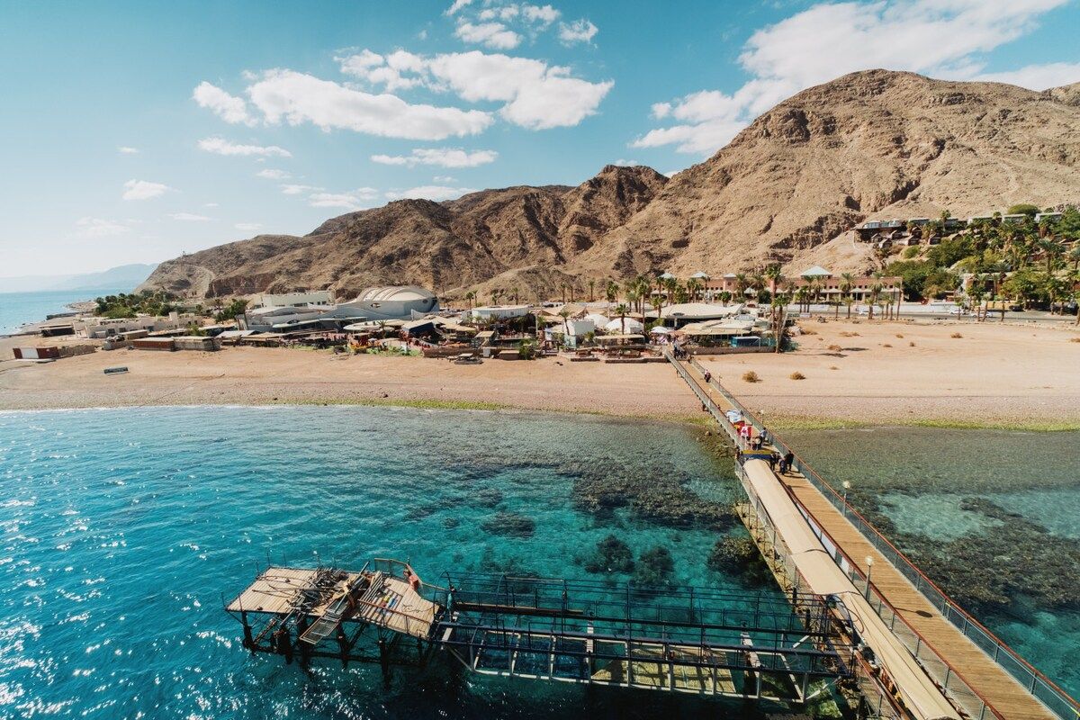 A coastal area with a pier extending into clear blue water, a beach with buildings, and a mountain in the background.