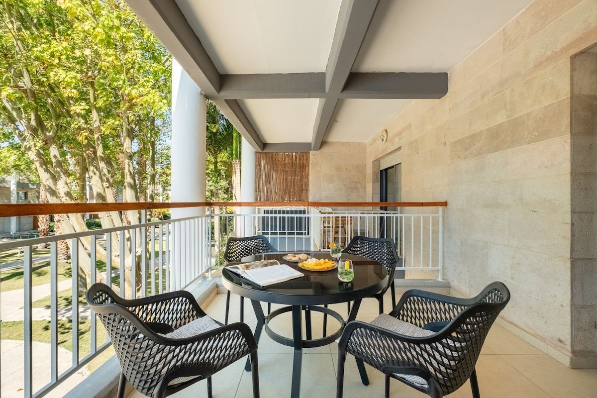 A balcony with a round table and four chairs, a wooden railing, and a view of trees and a building.