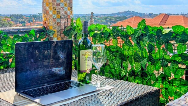Una computadora portátil sobre una mesa de mimbre con una botella y un vaso, un pilar de azulejos y vistas a follaje verde y edificios.
