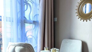 Una habitación con un sillón capitoné, una mesita redonda con mantel blanco y una silla blanca. Hay un espejo decorativo en la pared y una ventana con cortinas.