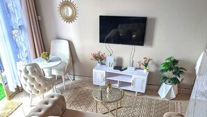 Una sala de estar con un sofá color beige, una mesa de centro de vidrio, una mesita auxiliar blanca, un televisor y una planta en maceta.