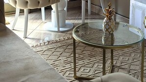 Una sala de estar con dos sillones blancos, una mesa de centro de vidrio con una piña como adorno, y un elemento decorativo en la pared con forma de sol.