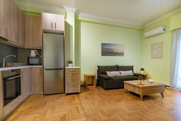 Una cocina moderna con refrigerador, horno y armarios, piso de madera en patrón de espiga, sofá, una mesita y un cuadro en la pared.