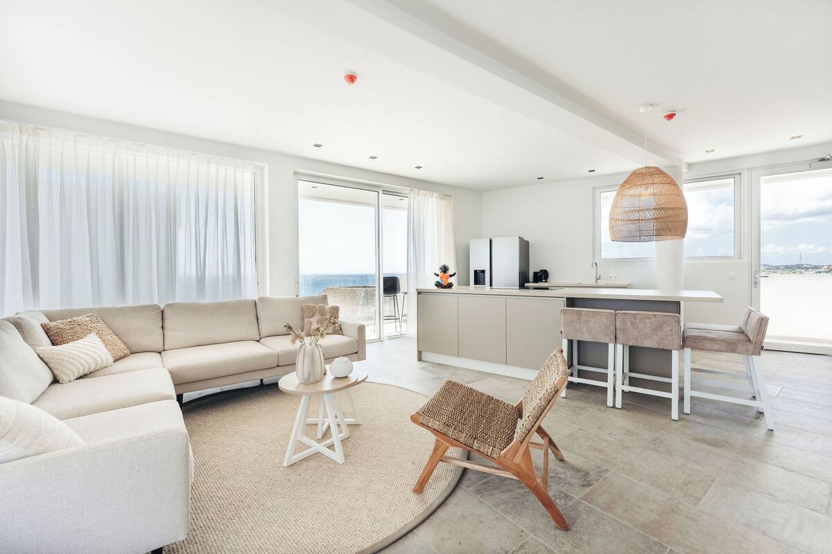 Ein modernes Wohnzimmer mit Sofa, rundem Tisch und Korbsessel. Es gibt eine Küchenzeile mit Bar und ein großes Fenster mit Blick auf das Meer.
