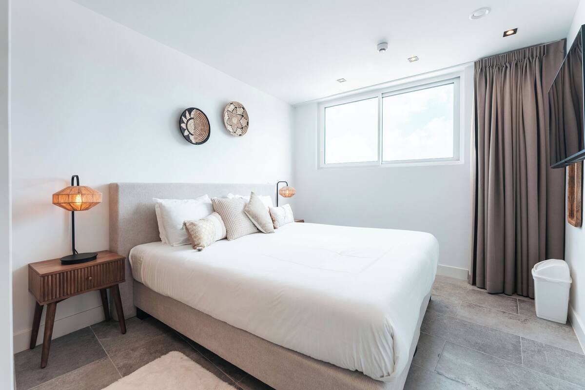 Ein Schlafzimmer mit einem großen Bett, zwei Nachttischen, einem Fenster mit Vorhängen und Wandbildern.
