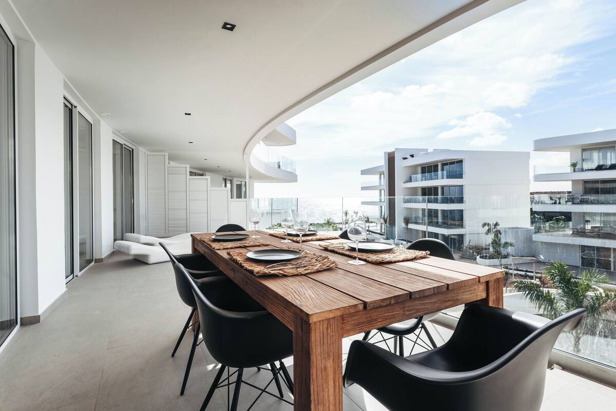 Ein moderner Essbereich mit einem Holztisch, gedeckt für sechs Personen, schwarzen Stühlen und Blick auf eine Stadtkulisse durch große Fenster.