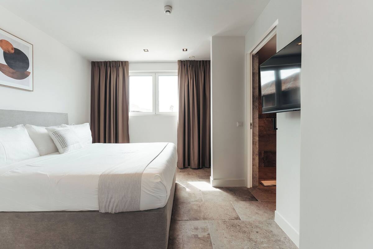 Ein modernes Hotelzimmer mit einem großen Bett, einem flachbildfernseher an der Wand und Blick ins Freie durch ein mit Vorhängen versehenes Fenster.