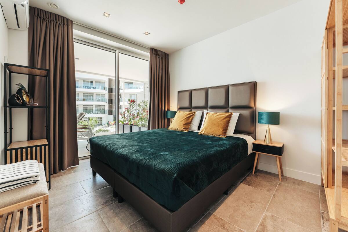 Ein modernes Schlafzimmer mit einem großen Bett, einem Nachttisch, einer Lampe und Blick auf den Balkon.