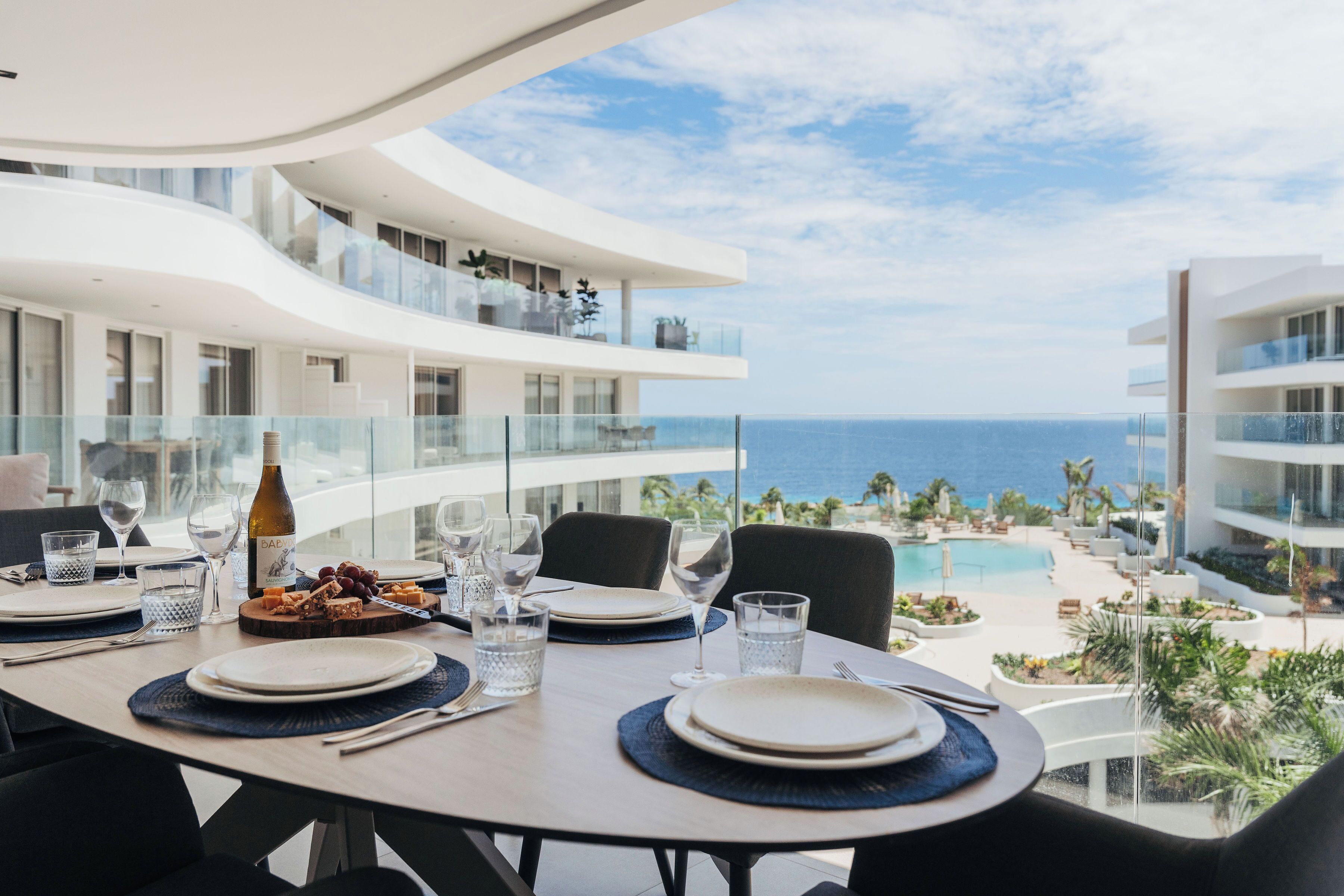Mesa de comedor dispuesta con platos, vasos y una botella, con vista a una piscina y al océano.