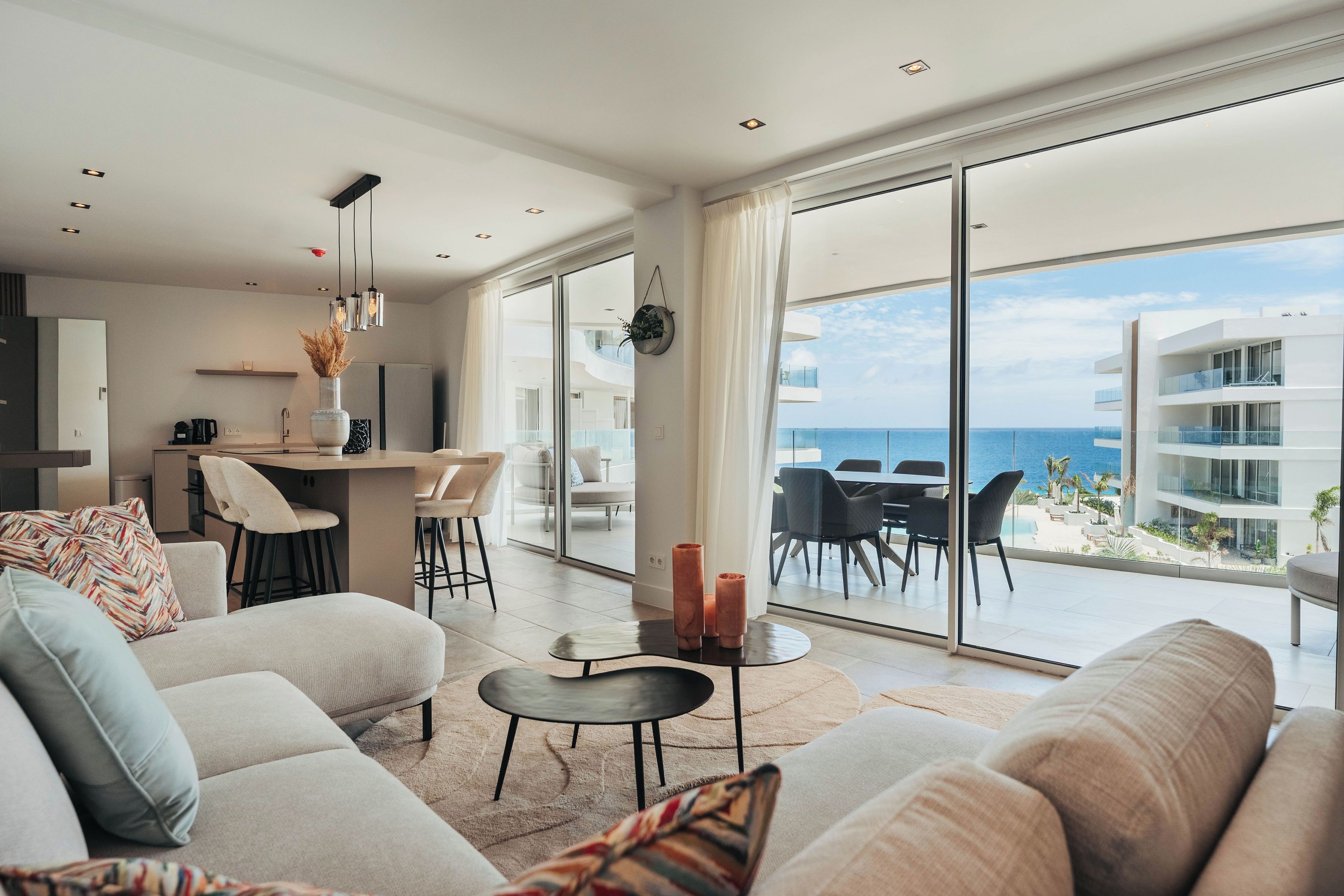 A modern living room with a sofa, coffee table, and a dining area with chairs and a table. Large windows offer a view of the ocean and a building across the street.