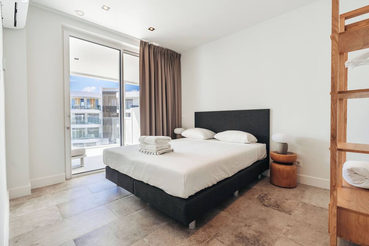 Ein modernes Schlafzimmer mit einem großen Bett, einem hölzernen Nachttisch und Blick auf die Stadt durch eine Schiebetür.