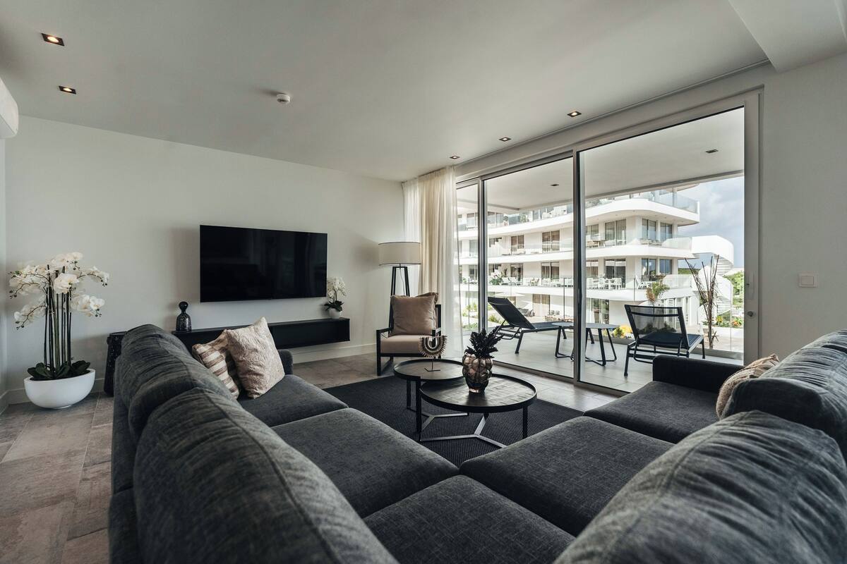 Ein modernes Wohnzimmer mit einer großen Couch, einem Flachbildfernseher und einem Balkon mit Blick auf ein Gebäude.