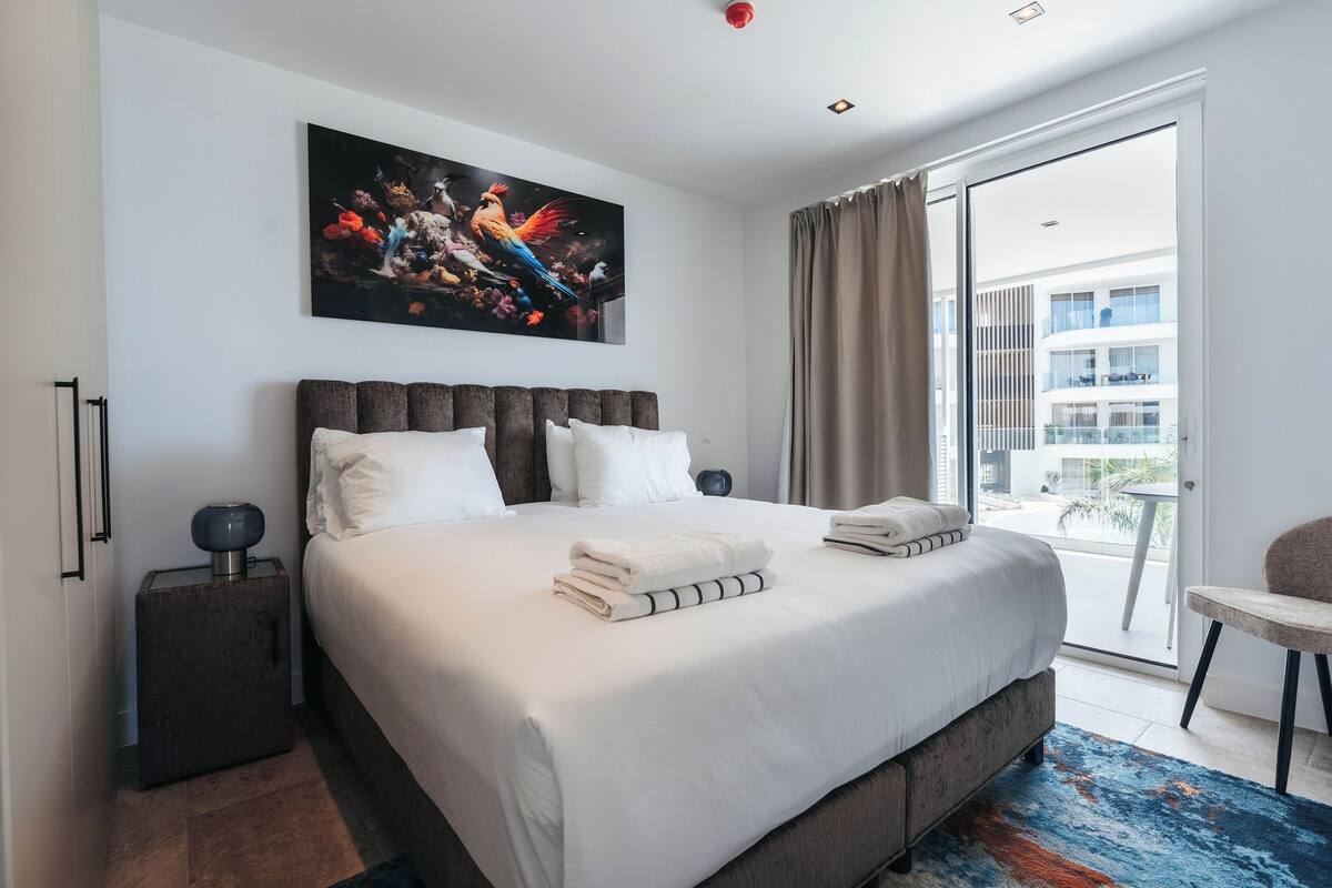 Ein modernes Hotelzimmer mit einem großen Bett, einem Bild darüber, einem Koffer und Blick auf den Balkon.