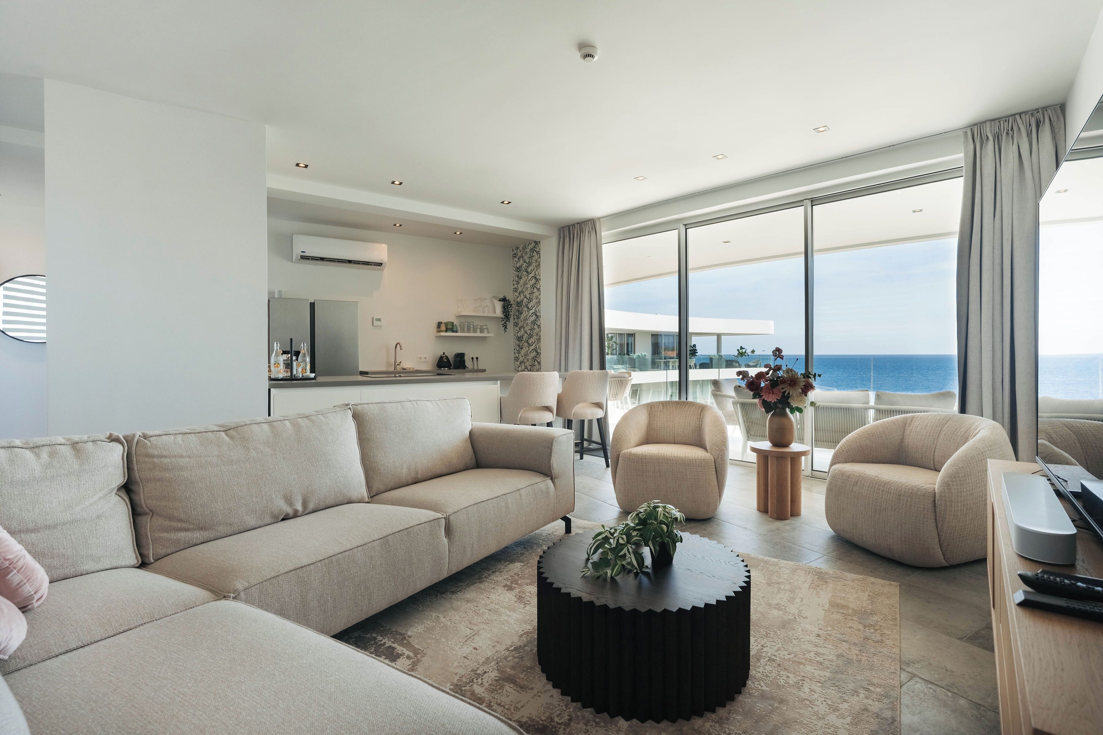 Un salon moderne avec un canapé beige, une table basse ronde et une vue sur l’océan grâce à de grandes fenêtres.