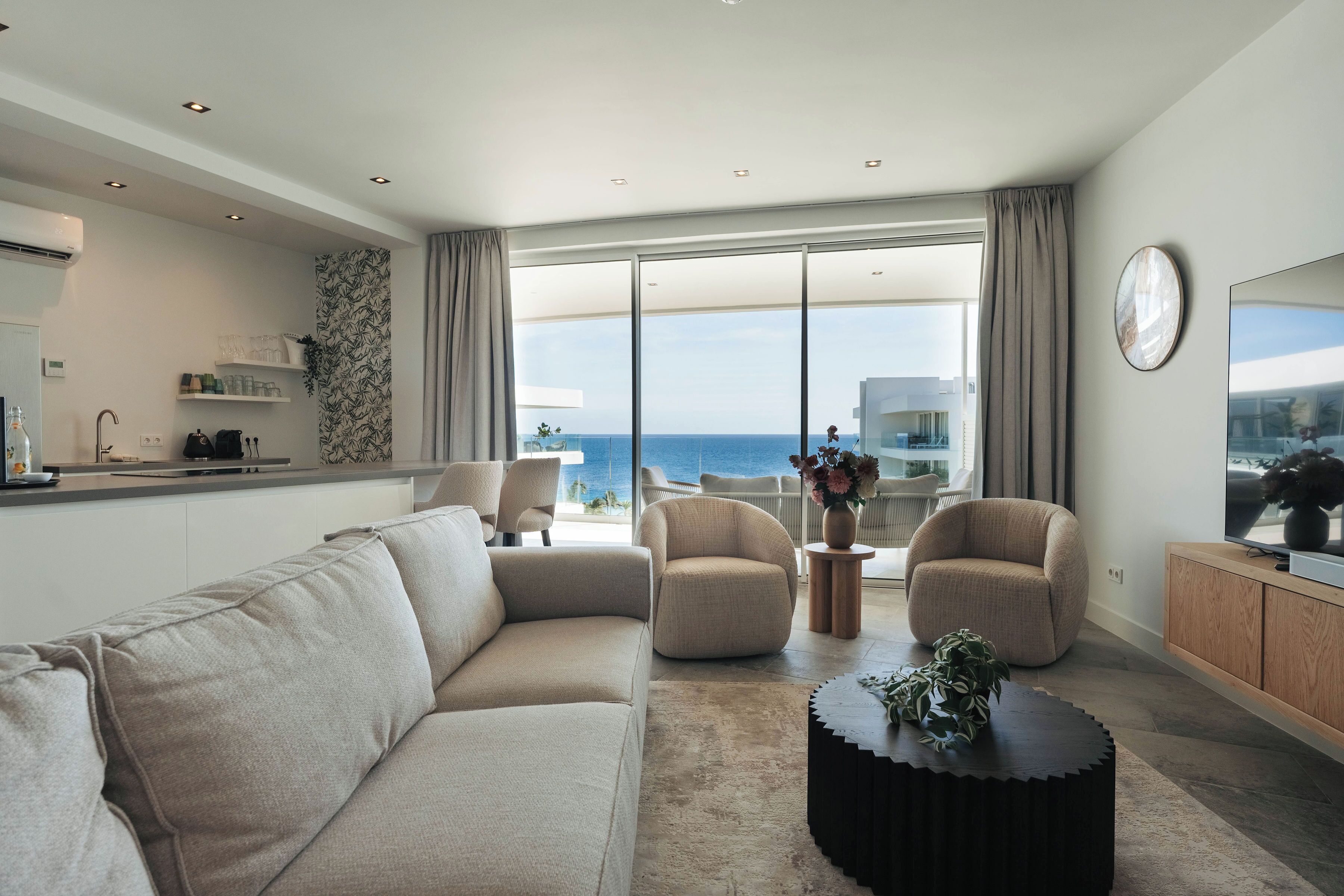 En moderne stue med en beige sofa, to lænestole, et sofabord og et fjernsyn på et træskab. Et stort vindue giver udsigt over havet.