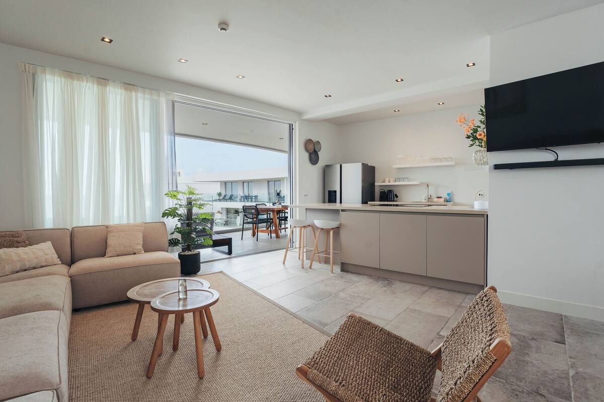 Ein modernes Wohnzimmer mit einer Couch, einem Couchtisch und einem Sessel. Es gibt eine Küchenzeile mit einem Kühlschrank, einem Fernseher und einem Esstisch mit Stühlen.