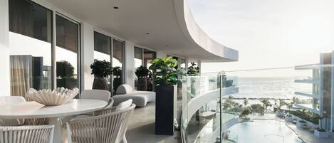 A modern balcony with a round table and chairs, potted plants, and a view of the ocean.
