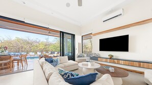 A modern living room with a large sofa, a coffee table, and a TV wall-mounted. There is a pool visible through the large windows. - 5BR Luxury Villa, Large Pool, 3-Min Walk to Beach, Concierge (Cabo Velas, Santa Cruz)