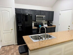 A compact kitchen with dark cabinets, a stainless steel sink, a black refrigerator, a microwave, and a coffee maker. - Poolside Paridise (New Braunfels)