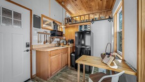 A compact kitchen with wooden cabinets, a black refrigerator, a microwave, and a small dining table with a chair. - Tiny Cub House | Secluded Tiny House on 1.7 Acres, Mentone (Mentone)