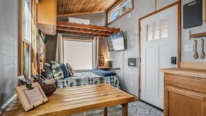A cozy room with a wooden table, a bed with a checkered blanket, a window, and a door. - Tiny Cub House | Secluded Tiny House on 1.7 Acres, Mentone (Mentone)