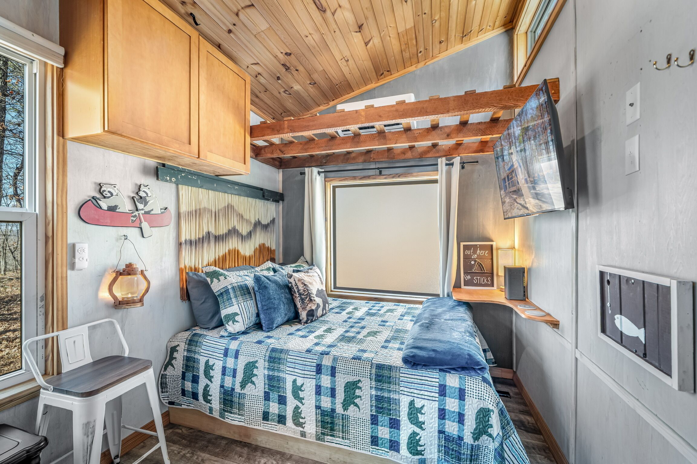 Un dormitorio acogedor de estilo cabaña, con techo de madera, una cama con colcha azul y blanca a cuadros, un escritorio pequeño y una ventana con vistas al exterior.