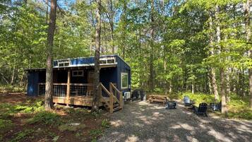 A small cabin with a deck and a covered area, surrounded by trees in a forest setting.