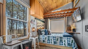 A cozy cabin interior with a bed, a small table, a chair, a fireplace, and large windows offering a forest view. - Tiny Cub House | Secluded Tiny House on 1.7 Acres, Mentone (Mentone)