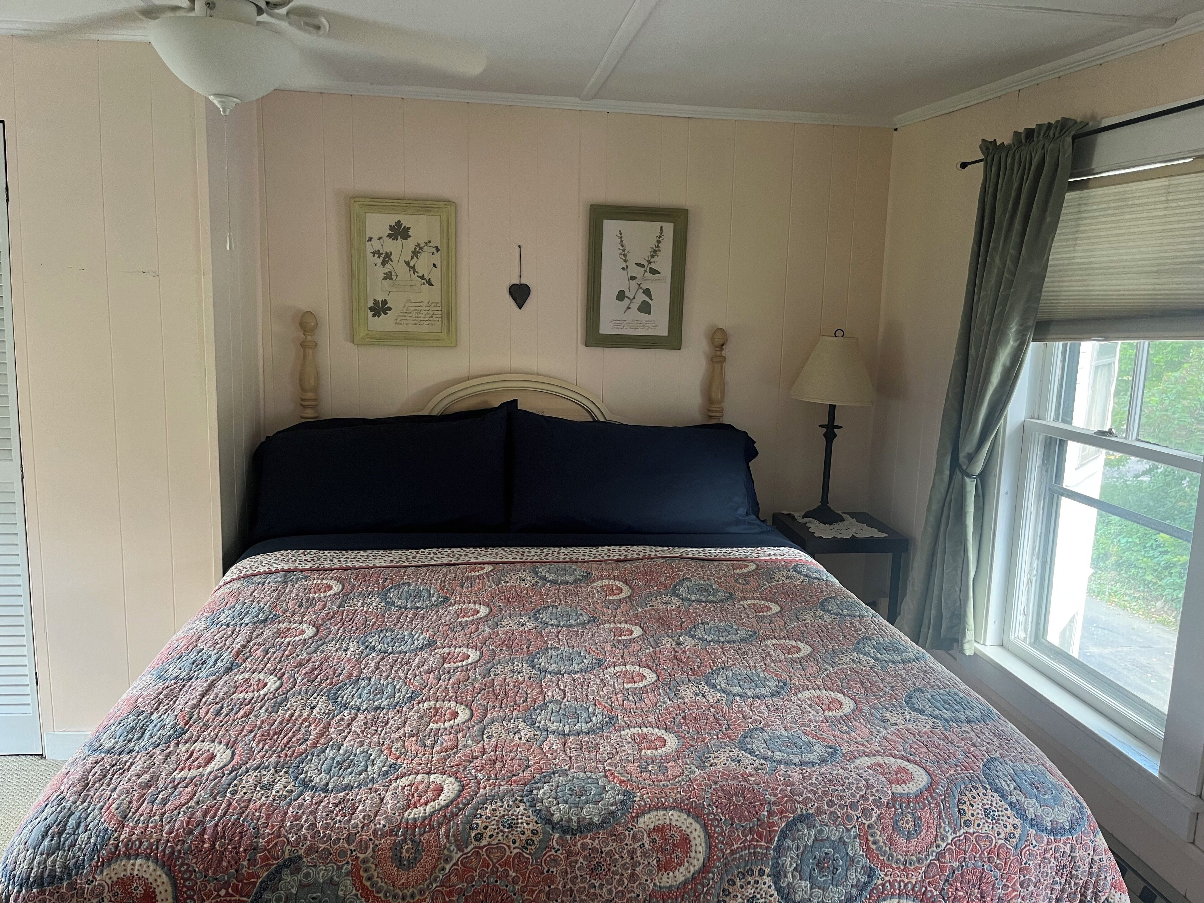 A bedroom with a large bed, a ceiling fan, a nightstand with a lamp, and a window with curtains.