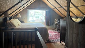 Un dormitorio acogedor en el ático con cama, dos lámparas de noche, una ventana con cortinas, un ventilador y un espejo.