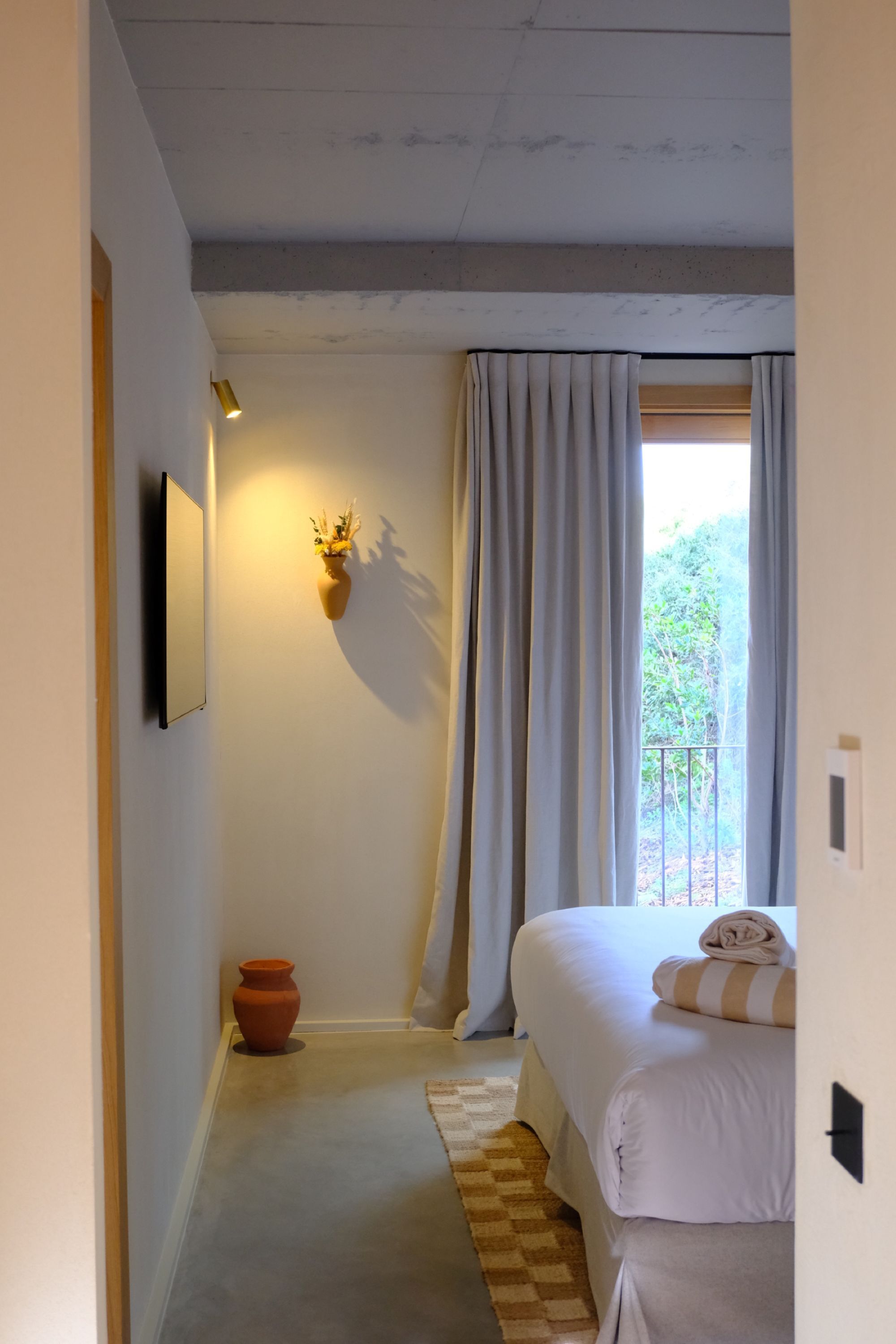 A bedroom with a bed, a wall-mounted light, a small potted plant, and a window with curtains.