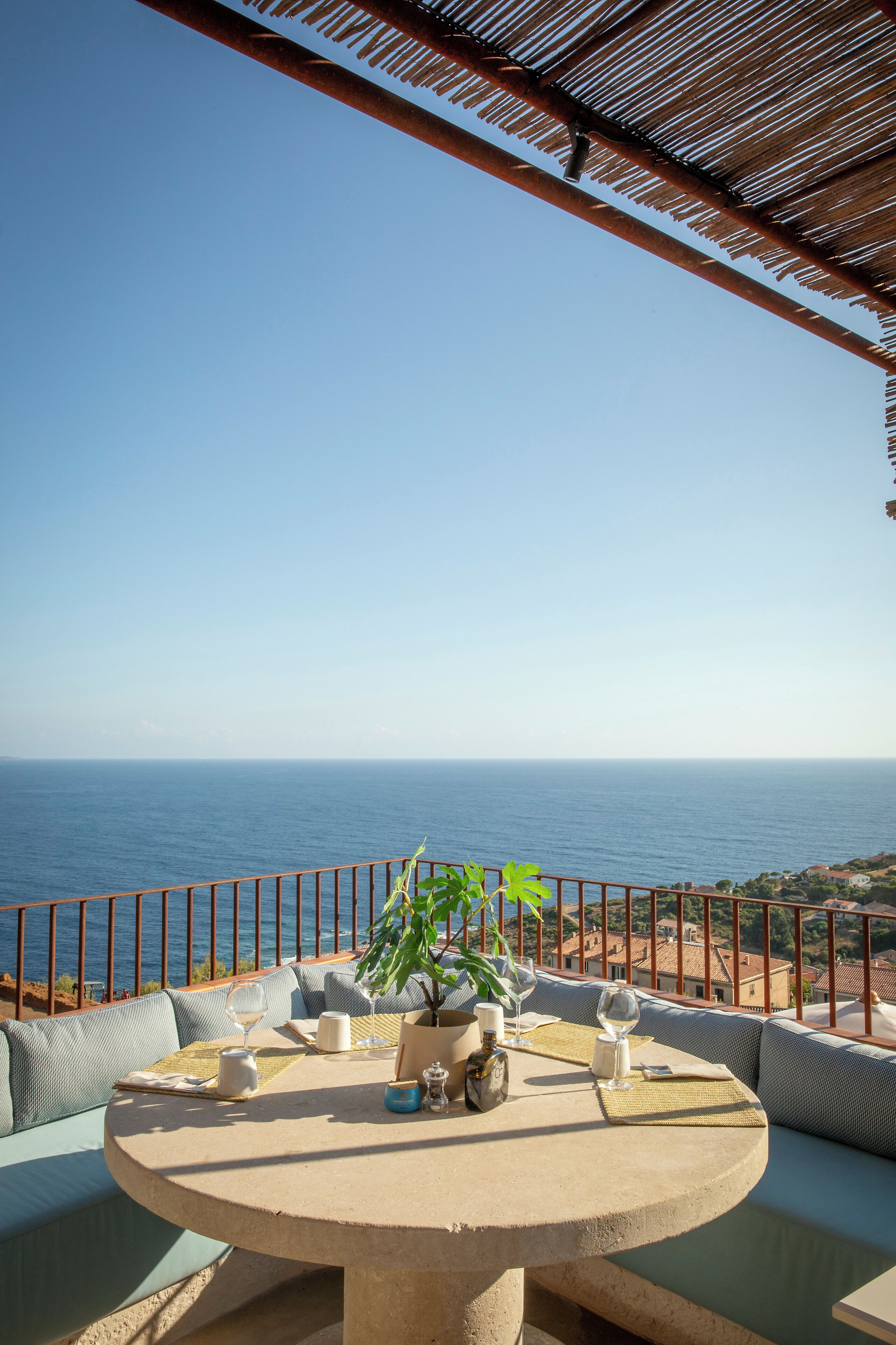 A round table with a view of the sea, a potted plant, and a glass of wine on a balcony with a railing.
