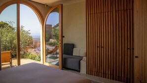 A bedroom with a large bed, a wooden wardrobe, and a sliding glass door leading to a balcony with a view of the sea. -  Hôtel Mylos - Cargèse (Cargèse)