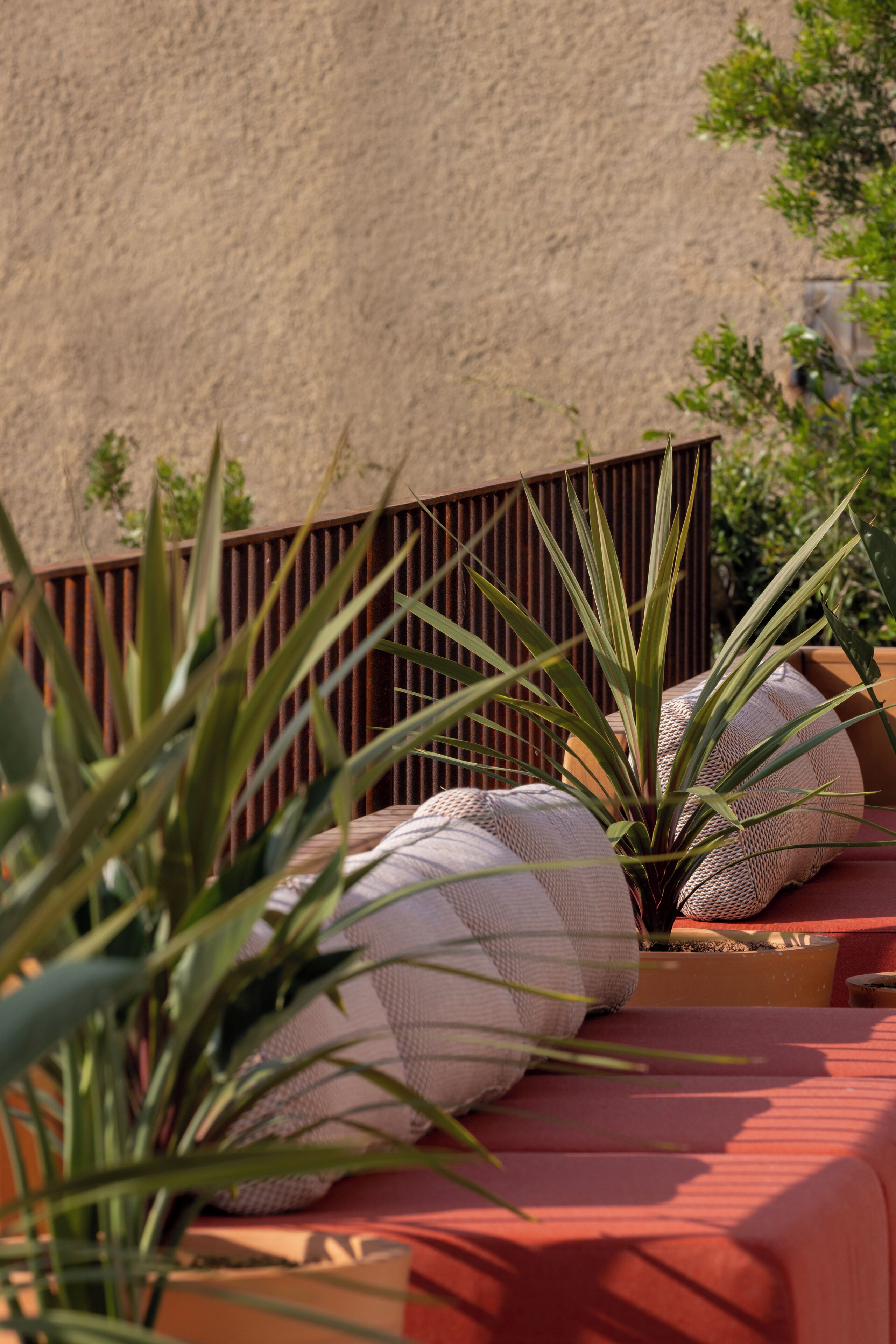 A sunlit outdoor seating area with a red cushioned bench, a potted plant, and a metal fence.