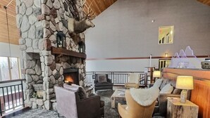 Une cheminée en pierre surmontée d’une tête de cerf, un coin salon avec des canapés en cuir, une table basse en bois et un mur en pierre.