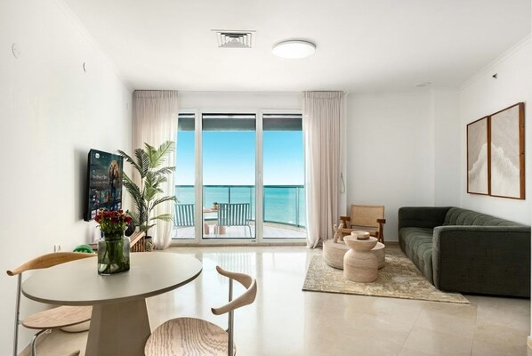 A modern living room with a sofa, a dining table with chairs, a coffee table, and a view of the sea through large windows.
