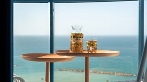 Une table avec deux verres contenant une boisson, une carafe et une vue sur l’océan par la fenêtre.